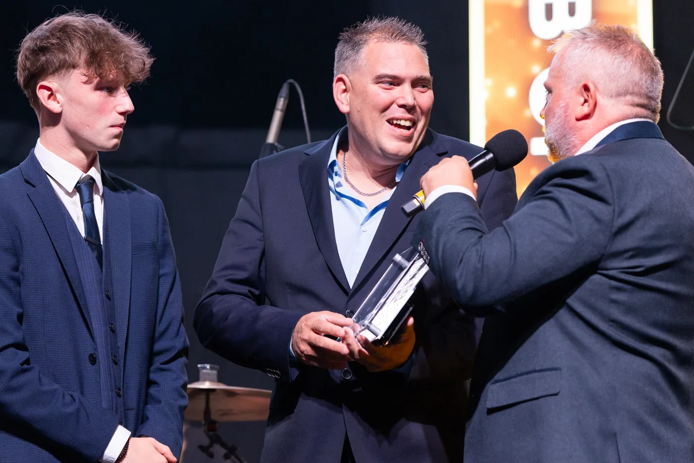 A member of The Honey Bee team holds the glass award on stage while being interviewed with a microphone, a younger team member beside him