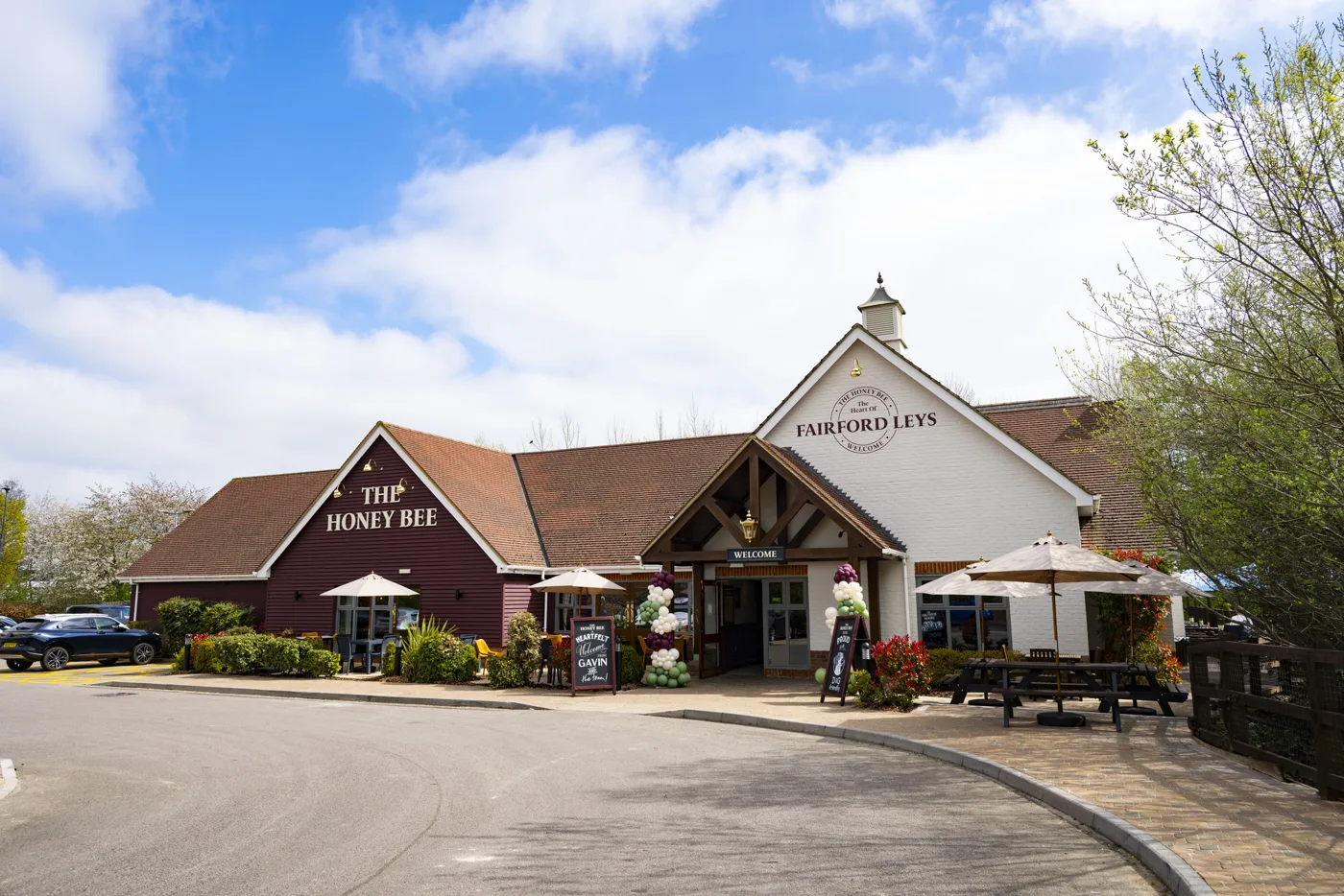 The Honey Bee pub from outside on a sunny day, a dark red gable with the pub's name beside a white gable reading Fairford Leys, with parasols and picnic benches out front