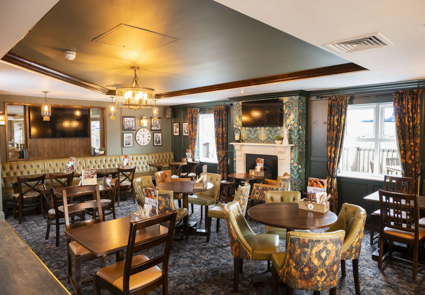 The lounge at The Honey Bee: round wooden tables, green leather and patterned chairs, a fireplace with a television above it and a long buttoned banquette